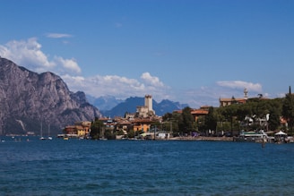 A scenic view of a lakeside town with a prominent castle tower amid charming, colorful buildings. The town is nestled beside a large, calm body of water with several boats. Majestic mountains provide a dramatic backdrop under a clear blue sky with scattered clouds.