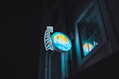 A neon sign attached to a building facade at night, featuring the words 'Rio Maravilha' with an adjacent arrow design. The sign is illuminated with blue and yellow colors and reflects in a nearby window, creating a vibrant glow against the dark background.