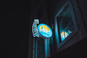 A neon sign attached to a building facade at night, featuring the words 'Rio Maravilha' with an adjacent arrow design. The sign is illuminated with blue and yellow colors and reflects in a nearby window, creating a vibrant glow against the dark background.