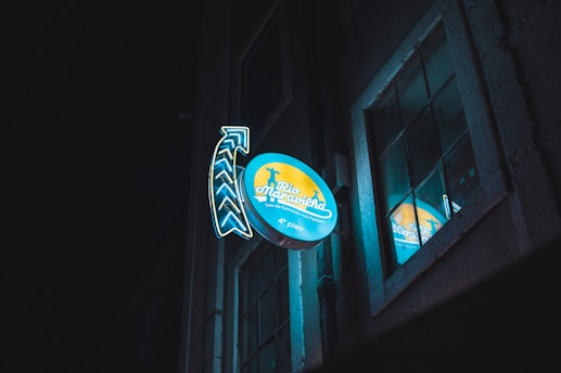 A neon sign attached to a building facade at night, featuring the words 'Rio Maravilha' with an adjacent arrow design. The sign is illuminated with blue and yellow colors and reflects in a nearby window, creating a vibrant glow against the dark background.