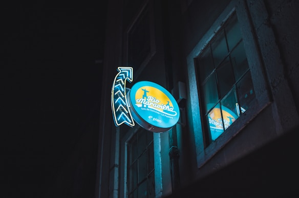 A neon sign attached to a building facade at night, featuring the words 'Rio Maravilha' with an adjacent arrow design. The sign is illuminated with blue and yellow colors and reflects in a nearby window, creating a vibrant glow against the dark background.