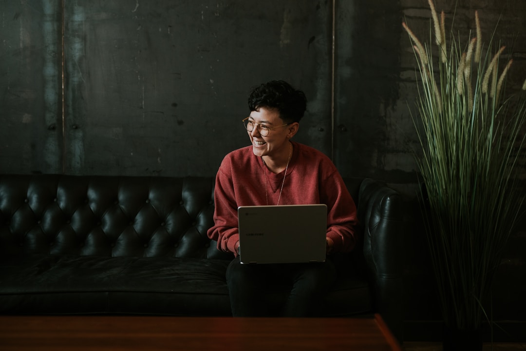 A person in a red sweater, laughing, while using their laptop and sitting on a black leather couch.