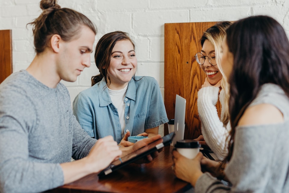 Four people sitting at a high table smiling with coffee, a laptop, and tablet,