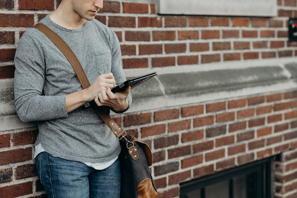 standing man in gray crew-neck long-sleeved shirt leaning on brown bricked wall