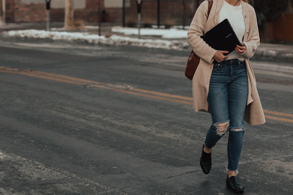 woman in beige coat and distressed blue jeans walking on road