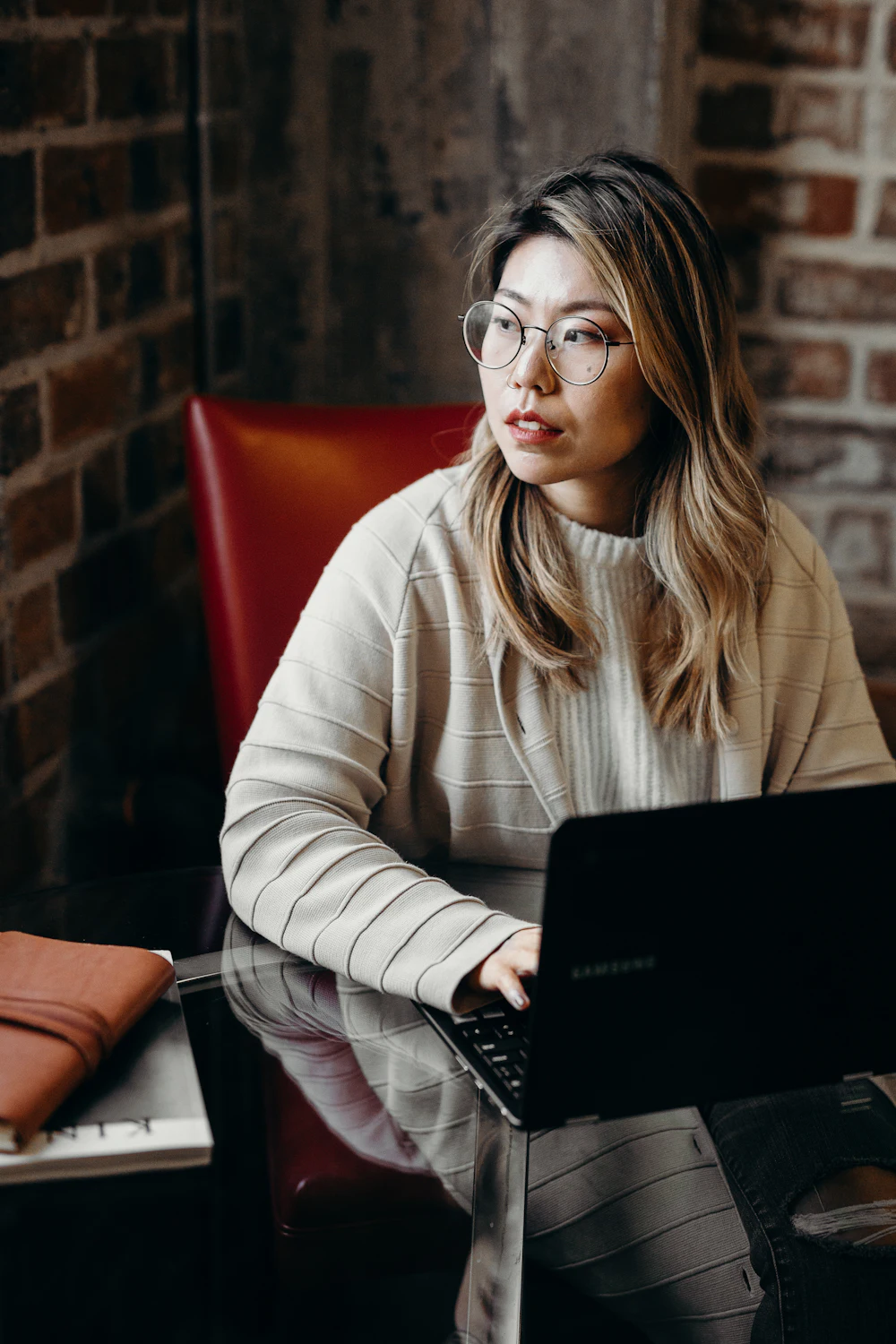A woman in round glasses and a cream knit working on her laptop at a cafe table
