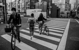 A black-and-white urban scene with people crossing a pedestrian crossing. Two people are pushing bicycles, and one person is cycling across. The background features city buildings and a quiet street, suggesting a typical city environment.