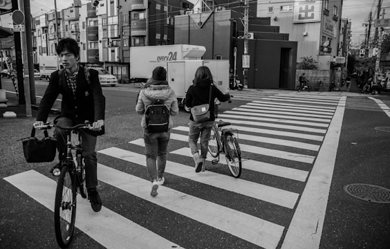A black-and-white urban scene with people crossing a pedestrian crossing. Two people are pushing bicycles, and one person is cycling across. The background features city buildings and a quiet street, suggesting a typical city environment.