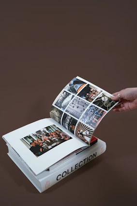 A hand is turning the page of an open photo album resting on a closed book titled 'Collection'. The album contains various photographs, including one of a group of people gathered around a dining table and several other images showcasing architectural details.