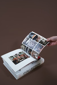 A hand is turning the page of an open photo album resting on a closed book titled 'Collection'. The album contains various photographs, including one of a group of people gathered around a dining table and several other images showcasing architectural details.