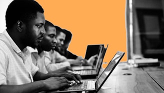 A group of people are focused on working with laptops, seated in a row at a wooden table. The background features an abstract orange shape contrasting with the monochrome setting of the foreground.