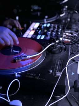 A DJ is operating a turntable with a red vinyl record. The setup includes a mixer board with various dials and glowing buttons. The environment appears to be dimly lit, suggesting a club or live performance setting.