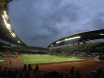 A large outdoor stadium with an illuminated field is depicted under a cloudy, evening sky. The stands hold scattered spectators, and the floodlights brighten the atmosphere. The image captures a panoramic view of a sports venue surrounded by a running track, with dark silhouettes of individuals in the foreground.