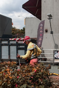 A worker dressed in a yellow high-visibility vest and red cap is standing near a metal container. The scene includes a building wall with a mounted camera and nearby plants with red flowers. It appears to be an urban setting.