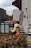 A worker dressed in a yellow high-visibility vest and red cap is standing near a metal container. The scene includes a building wall with a mounted camera and nearby plants with red flowers. It appears to be an urban setting.