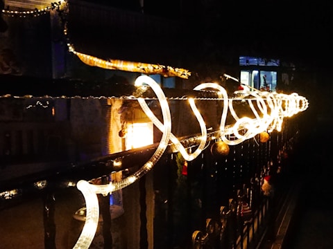 A series of bright, intertwined lights adorn a dark railing, creating a festive and warm atmosphere. In the background, a softly lit interior is visible through a window, adding to the cozy ambiance. Decorations hang along the railing, enhancing the scene's festive charm.