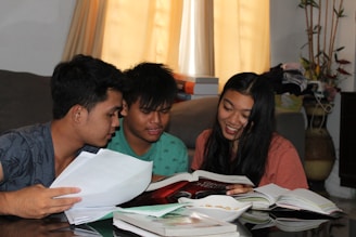 Three young people are sitting together indoors, engaging with books and papers. They appear to be studying or discussing something in a relaxed atmosphere. The room has a couch, books, and a potted plant in the background.