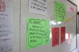 A classroom whiteboard with colorful educational posters and notes. Each poster contains scientific text, with some featuring illustrations or diagrams. Notes are written in various colors, including green, red, and pink papers pinned to the board. A hand is partially visible, suggesting interaction with the board.