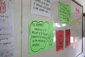 A classroom whiteboard with colorful educational posters and notes. Each poster contains scientific text, with some featuring illustrations or diagrams. Notes are written in various colors, including green, red, and pink papers pinned to the board. A hand is partially visible, suggesting interaction with the board.
