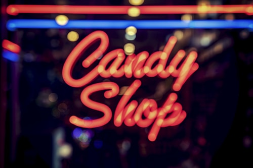 Neon red letters display the words 'Candy Shop' against a blurred, dark background. Blue and red lines frame the text, suggesting a vibrant, nighttime urban setting.