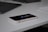 A smartphone rests on a plain white surface next to a sleek black laptop and part of a white keyboard. The smartphone's screen reflects icons and applications, indicating it is on a desk setup likely used for work or study.