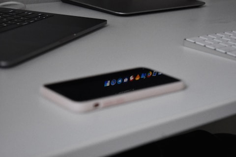 A smartphone rests on a plain white surface next to a sleek black laptop and part of a white keyboard. The smartphone's screen reflects icons and applications, indicating it is on a desk setup likely used for work or study.