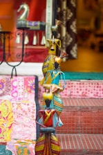 Colorful handcrafted figures, each adorned with vibrant and intricate patterns, are arranged on a set of tiled steps. A decorative horse statue and embroidered textiles with floral motifs add to the eclectic display, creating an artistic and cultural ambiance.