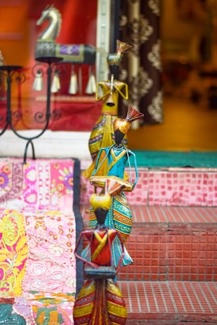 Colorful handcrafted figures, each adorned with vibrant and intricate patterns, are arranged on a set of tiled steps. A decorative horse statue and embroidered textiles with floral motifs add to the eclectic display, creating an artistic and cultural ambiance.