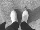 A pair of light-colored sneakers are visible against a textured, rough surface, suggesting a concrete or asphalt ground. The sneakers have a knitted upper material with visible laces and a clean, casual style. Partial shadow of an individual is visible due to the angle of sunlight.