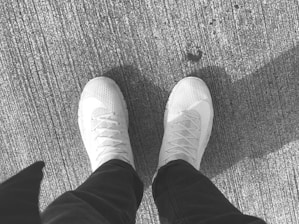 A pair of light-colored sneakers are visible against a textured, rough surface, suggesting a concrete or asphalt ground. The sneakers have a knitted upper material with visible laces and a clean, casual style. Partial shadow of an individual is visible due to the angle of sunlight.
