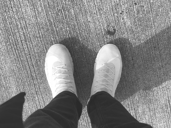 A pair of light-colored sneakers are visible against a textured, rough surface, suggesting a concrete or asphalt ground. The sneakers have a knitted upper material with visible laces and a clean, casual style. Partial shadow of an individual is visible due to the angle of sunlight.