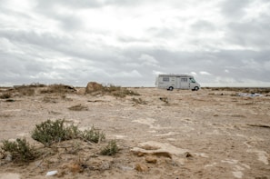 A recreational vehicle is parked on a barren, rocky landscape. The ground is covered with sparse bushes and the sky is overcast with grey clouds. The horizon appears vast and endless, contributing to a sense of solitude.