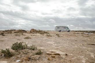 A recreational vehicle is parked on a barren, rocky landscape. The ground is covered with sparse bushes and the sky is overcast with grey clouds. The horizon appears vast and endless, contributing to a sense of solitude.