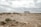 A recreational vehicle is parked on a barren, rocky landscape. The ground is covered with sparse bushes and the sky is overcast with grey clouds. The horizon appears vast and endless, contributing to a sense of solitude.