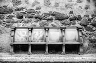 An intricately carved bench with detailed wooden panels set against a rough stone wall. The bench consists of four sections, each featuring decorative elements and sturdy legs.