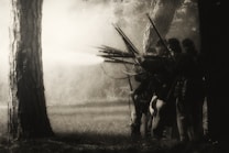 A group of soldiers dressed in historical military uniforms is positioned behind trees, aiming long rifles. Smoke is visible, suggesting the firing of weapons. The image is in sepia tones, giving it a historical or vintage feel.