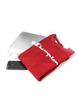 A neatly folded red sweatshirt with the word 'Champion' printed on it in white, placed on top of a closed silver laptop. Nearby are a black smartphone and a pair of white wireless earbuds, all set against a clean white background.