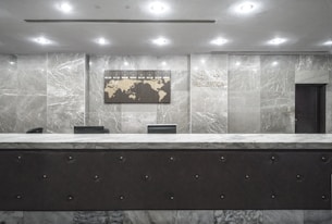 An empty reception desk with a marble finish is situated against a backdrop of polished stone walls. A decorative wall map and signage labeled 'Reception' are mounted above the desk. The lighting is soft and evenly distributed across the room.