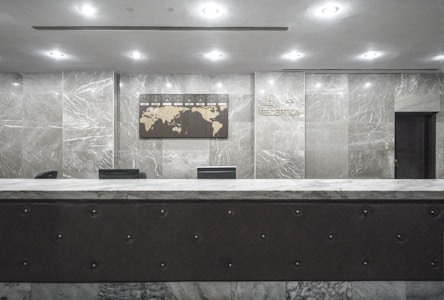 An empty reception desk with a marble finish is situated against a backdrop of polished stone walls. A decorative wall map and signage labeled 'Reception' are mounted above the desk. The lighting is soft and evenly distributed across the room.