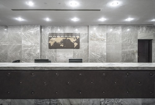 An empty reception desk with a marble finish is situated against a backdrop of polished stone walls. A decorative wall map and signage labeled 'Reception' are mounted above the desk. The lighting is soft and evenly distributed across the room.
