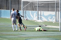 Two soccer players are on a field near the goalpost. One player is wearing a white jersey with the number 4 and walking away from the goal. Another player in a dark jersey appears to be celebrating a goal. A goalkeeper wearing a bright green uniform is lying on the ground.