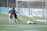 Two soccer players are on a field near the goalpost. One player is wearing a white jersey with the number 4 and walking away from the goal. Another player in a dark jersey appears to be celebrating a goal. A goalkeeper wearing a bright green uniform is lying on the ground.