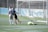 Two soccer players are on a field near the goalpost. One player is wearing a white jersey with the number 4 and walking away from the goal. Another player in a dark jersey appears to be celebrating a goal. A goalkeeper wearing a bright green uniform is lying on the ground.