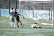 Two soccer players are on a field near the goalpost. One player is wearing a white jersey with the number 4 and walking away from the goal. Another player in a dark jersey appears to be celebrating a goal. A goalkeeper wearing a bright green uniform is lying on the ground.