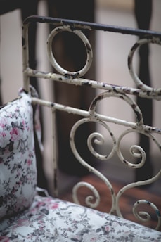 An ornate, vintage-style metal headboard with circular and swirl designs, next to a floral-patterned pillow. The pillow features pink and grey flowers on a white background, creating a shabby chic aesthetic.