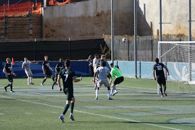 A soccer game is in progress with players from two teams, one in white uniforms and the other in dark green uniforms. The ball is in mid-air near the goal, with the goalkeeper in a green jersey preparing to make a save. Several players are positioned around the goal area, and a netted fence is visible in the background.