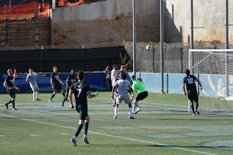 A soccer game is in progress with players from two teams, one in white uniforms and the other in dark green uniforms. The ball is in mid-air near the goal, with the goalkeeper in a green jersey preparing to make a save. Several players are positioned around the goal area, and a netted fence is visible in the background.