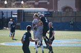 A group of soccer players is actively competing for the ball in the air, showcasing the intensity of the match. The field has bright green artificial turf, and the backdrop includes some blurred buildings and fencing. The players are wearing uniforms with numbers, indicating an organized game, and the sunlight casts sharp shadows on the ground.