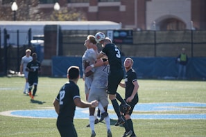 A group of soccer players is actively competing for the ball in the air, showcasing the intensity of the match. The field has bright green artificial turf, and the backdrop includes some blurred buildings and fencing. The players are wearing uniforms with numbers, indicating an organized game, and the sunlight casts sharp shadows on the ground.