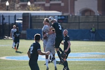 A group of soccer players is actively competing for the ball in the air, showcasing the intensity of the match. The field has bright green artificial turf, and the backdrop includes some blurred buildings and fencing. The players are wearing uniforms with numbers, indicating an organized game, and the sunlight casts sharp shadows on the ground.